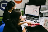 Two individuals are engaged in a robotics task at a desk equipped with a computer displaying a 3D model of a robotic vehicle. They are surrounded by various robotic parts and kits, suggesting involvement in a constructive or educational activity.
