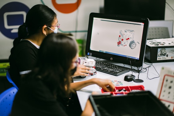 Two individuals are engaged in a robotics task at a desk equipped with a computer displaying a 3D model of a robotic vehicle. They are surrounded by various robotic parts and kits, suggesting involvement in a constructive or educational activity.
