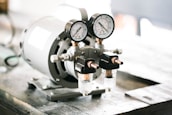 A pressure gauge mounted on a piece of industrial equipment with two glass components attached below it, likely indicating the presence of a pneumatic or hydraulic system. The equipment rests on a wooden surface.