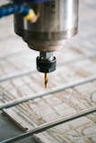 A close-up view of a CNC machine with a drill bit poised above a sheet of material partially carved with intricate patterns. The metal spindle holding the drill is prominently in focus, with the surrounding environment slightly blurred.