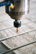 A close-up view of a CNC machine with a drill bit poised above a sheet of material partially carved with intricate patterns. The metal spindle holding the drill is prominently in focus, with the surrounding environment slightly blurred.