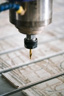 A close-up view of a CNC machine with a drill bit poised above a sheet of material partially carved with intricate patterns. The metal spindle holding the drill is prominently in focus, with the surrounding environment slightly blurred.