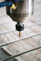 A close-up view of a CNC machine with a drill bit poised above a sheet of material partially carved with intricate patterns. The metal spindle holding the drill is prominently in focus, with the surrounding environment slightly blurred.