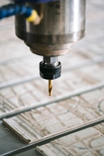 A close-up view of a CNC machine with a drill bit poised above a sheet of material partially carved with intricate patterns. The metal spindle holding the drill is prominently in focus, with the surrounding environment slightly blurred.