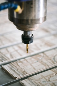 A close-up view of a CNC machine with a drill bit poised above a sheet of material partially carved with intricate patterns. The metal spindle holding the drill is prominently in focus, with the surrounding environment slightly blurred.