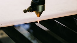 A laser cutting machine is in the process of cutting through a sheet of material. The laser emits a bright light as it precisely cuts along the edge. The machine's nozzle is positioned closely to the material, demonstrating a high level of precision.