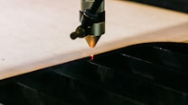 A laser cutter is in operation, with a beam slicing through a sheet of material placed on a cutting bed. The metallic nozzle of the machine is visible, directing the laser precisely along the edge.