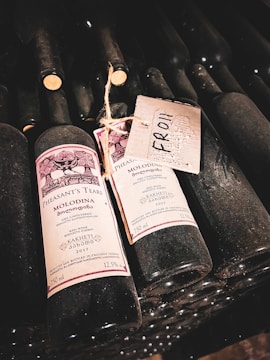 Several wine bottles are stacked in a wine cellar, with two prominently displayed in the foreground. The labels read 'Pheasant's Tears Molodina', indicating the wine's origin and vintage as 2017. A handwritten tag labeled 'From' is attached to one bottle with twine.