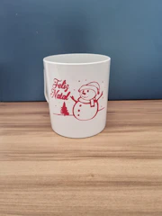 A white mug featuring a red line drawing of a snowman, wearing a hat and scarf, next to a pine tree. The mug also has the words 'Feliz Natal' in red cursive text. The scene is set against a wooden surface with a plain, dark background.