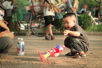 A young boy squats on the ground, intently playing with a colorful toy robot. Nearby, two bottles are placed on the pavement, next to what appears to be an adult's knee. In the background, there are other people walking, bicycles, and trees, suggesting a park setting.