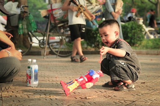 A young boy squats on the ground, intently playing with a colorful toy robot. Nearby, two bottles are placed on the pavement, next to what appears to be an adult's knee. In the background, there are other people walking, bicycles, and trees, suggesting a park setting.