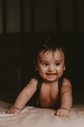 A smiling baby is lying on their stomach on a bed. The baby has dark hair and is wearing a simple black garment. The background is dimly lit, creating a warm and intimate atmosphere.