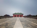 A large, historic Chinese temple stands at the end of a wide, paved pathway. The building features traditional architecture with a green-tiled roof and red-walled structure. Flanked by two smaller buildings, it is surrounded by a black metal fence. The sky is overcast, creating a calm ambiance.