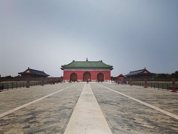 A large, historic Chinese temple stands at the end of a wide, paved pathway. The building features traditional architecture with a green-tiled roof and red-walled structure. Flanked by two smaller buildings, it is surrounded by a black metal fence. The sky is overcast, creating a calm ambiance.