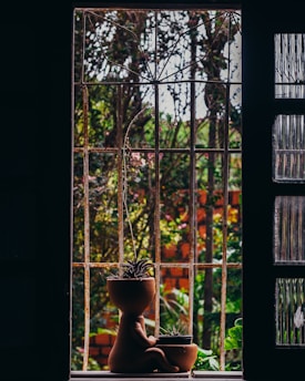 A window with metal bars overlooks a garden, with two clay pots shaped like human figures placed on the windowsill. Each pot contains a small plant, and the background is filled with greenery and a brick wall.
