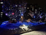 A snowy landscape is illuminated by colorful string lights wrapped around trees and bushes. The lights cast a warm, festive glow with bright blue, red, and gold hues. A small lantern positioned on the ground enhances the enchanting atmosphere. In the background, residential buildings add depth with their lit windows visible against the night sky.