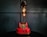 A vintage-style red rotary telephone is creatively repurposed into a lamp. The lamp features two exposed Edison bulbs mounted on a sleek black support extending from the phone's base. The background consists of a dark, muted wall with subtle paneling, which enhances the industrial and retro aesthetic.