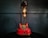 A vintage-style red rotary telephone is creatively repurposed into a lamp. The lamp features two exposed Edison bulbs mounted on a sleek black support extending from the phone's base. The background consists of a dark, muted wall with subtle paneling, which enhances the industrial and retro aesthetic.