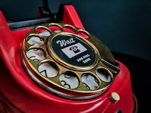 A close-up of a vintage red rotary telephone with a gold dial that has numbers arranged in a circular fashion. The central part of the dial features a sticker with the words 'Wait for dial tone' and an image of a telephone handset.
