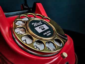 A close-up of a vintage red rotary telephone with a gold dial that has numbers arranged in a circular fashion. The central part of the dial features a sticker with the words 'Wait for dial tone' and an image of a telephone handset.