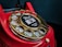 A close-up of a vintage red rotary telephone with a gold dial that has numbers arranged in a circular fashion. The central part of the dial features a sticker with the words 'Wait for dial tone' and an image of a telephone handset.