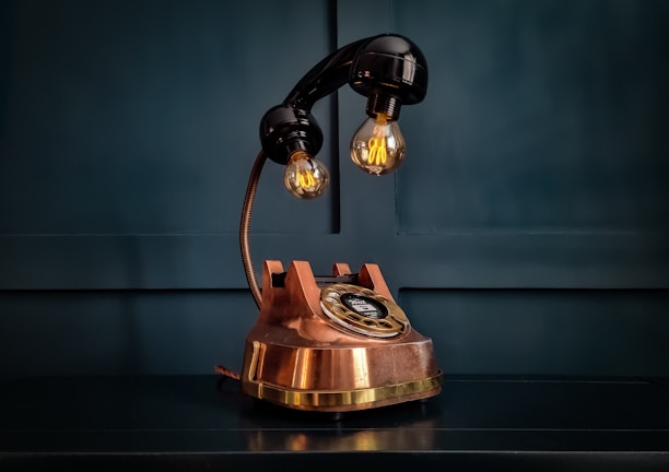 A vintage-style rotary telephone transformed into a lamp with two exposed bulbs, sitting on a dark surface. The telephone's base is copper with a black handset, contributing to its antique appearance. The background is a deep teal color, adding contrast.