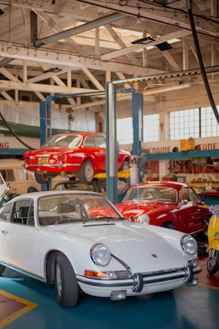 A vintage car workshop interior with several classic cars. A white vintage sports car is parked on the ground, while a red car is elevated on a car lift in the background. Another red vehicle and a yellow scooter are parked nearby. The workshop has a rustic look with wooden beams and large windows letting in natural light.