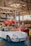 A vintage car workshop interior with several classic cars. A white vintage sports car is parked on the ground, while a red car is elevated on a car lift in the background. Another red vehicle and a yellow scooter are parked nearby. The workshop has a rustic look with wooden beams and large windows letting in natural light.