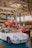 A vintage car workshop interior with several classic cars. A white vintage sports car is parked on the ground, while a red car is elevated on a car lift in the background. Another red vehicle and a yellow scooter are parked nearby. The workshop has a rustic look with wooden beams and large windows letting in natural light.