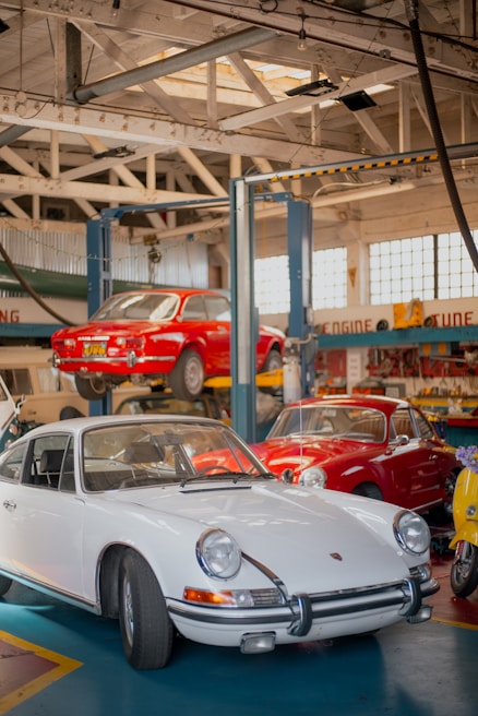 A vintage car workshop interior with several classic cars. A white vintage sports car is parked on the ground, while a red car is elevated on a car lift in the background. Another red vehicle and a yellow scooter are parked nearby. The workshop has a rustic look with wooden beams and large windows letting in natural light.