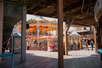 Underneath a wooden boardwalk, a brightly colored carousel with intricate gold details stands surrounded by small stalls and people. Some individuals are walking, while others are engaged with the attractions. The atmosphere is vibrant and lively, typical of a fair or amusement park setting.