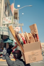 A person carries a large cardboard box filled with various smaller boxes and packaging materials on their shoulder, walking along a city street. The street is lined with buildings and a street sign is visible in the background.