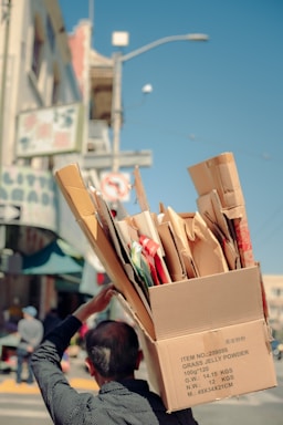 A person carries a large cardboard box filled with various smaller boxes and packaging materials on their shoulder, walking along a city street. The street is lined with buildings and a street sign is visible in the background.