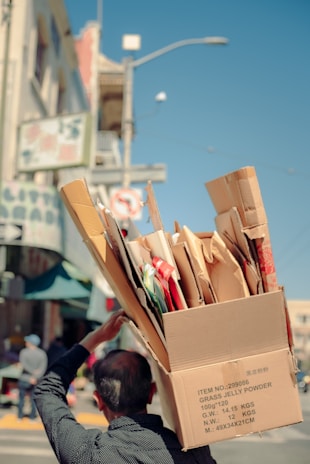A person carries a large cardboard box filled with various smaller boxes and packaging materials on their shoulder, walking along a city street. The street is lined with buildings and a street sign is visible in the background.