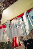 Several graphic sweatshirts are hanging on display outdoors. The clothes have various designs and logos, with some referencing San Francisco. They are arranged in a row under a bright yellow awning.