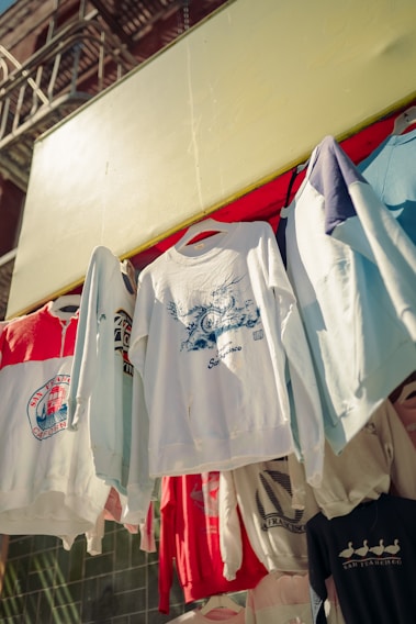 Several graphic sweatshirts are hanging on display outdoors. The clothes have various designs and logos, with some referencing San Francisco. They are arranged in a row under a bright yellow awning.