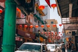A vibrant street scene in a Chinatown area featuring numerous red lanterns hanging above a busy street. Traditional Chinese architecture with colorful facades lines the sides, and various shops and restaurants are visible. People are walking along the sidewalks, and cars are parked along the street. Signs in both English and Chinese are prominently displayed.