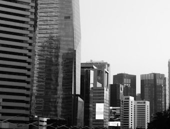 Tall skyscrapers with modern architectural designs dominate the cityscape. The buildings are made of glass and steel, with reflective surfaces creating a dynamic visual effect. Corporate logos are visible on some of the structures, indicating a business district. The image is in black and white, highlighting the contrast between light and shadow.