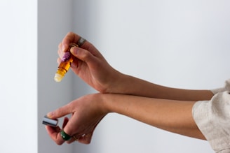 A person is applying an essential oil roller to their forearm. The bottle is amber-colored and has a label. The person is holding the cap in their other hand, and they are wearing rings.