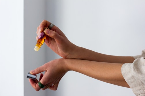 A person is applying an essential oil roller to their forearm. The bottle is amber-colored and has a label. The person is holding the cap in their other hand, and they are wearing rings.