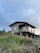 A wooden house on stilts is situated in a lush, green environment. The house has a simple structure and is elevated above the ground, supported by wooden poles. The surrounding area is filled with tall grass, vibrant orange flowers, and dense vegetation. The sky is overcast with a soft, moody light.