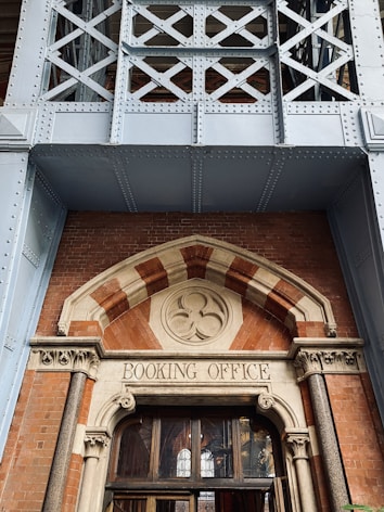 The entrance of a building showcases an ornate brick and stone facade with a decorative arch above a wooden door. The words 'Booking Office' are carved into a stone panel above the door, which is framed by intricate stone and brickwork. Above the entrance is a detailed three-leaf clover design, adding an element of historical architecture. The structure is further reinforced by a large steel framework with criss-cross patterns, giving it an industrial aesthetic.