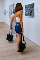 Two women stand in an art gallery observing framed pictures on a white wall. The woman closest to the camera has wavy brown hair and is dressed in a white top and denim shorts, holding a black bag. The floor is wooden, contributing to a clean and minimalist atmosphere.