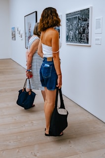 Two women stand in an art gallery observing framed pictures on a white wall. The woman closest to the camera has wavy brown hair and is dressed in a white top and denim shorts, holding a black bag. The floor is wooden, contributing to a clean and minimalist atmosphere.