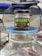 A jar of organic Black Forest honey sits on top of a large glass container with a blue lid. The jar has a black lid and features bold green and yellow labels with text in English and Arabic.
