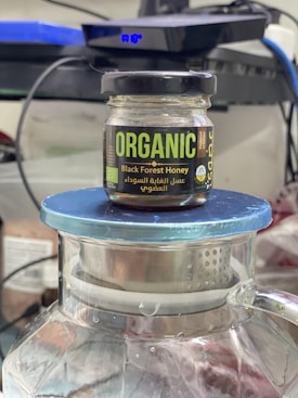 A jar of organic Black Forest honey sits on top of a large glass container with a blue lid. The jar has a black lid and features bold green and yellow labels with text in English and Arabic.
