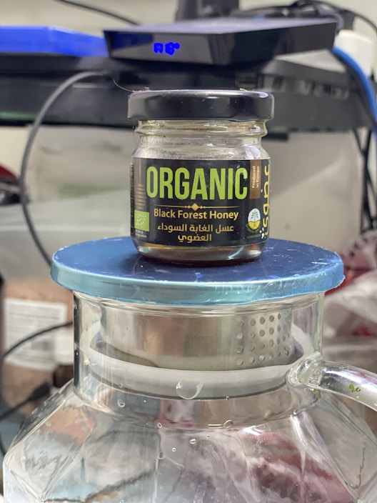 A jar of organic Black Forest honey sits on top of a large glass container with a blue lid. The jar has a black lid and features bold green and yellow labels with text in English and Arabic.