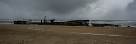 A rusted and partially submerged shipwreck lies on a sandy beach under a cloudy, overcast sky. The dark, looming clouds add to the somber atmosphere, while the ocean waves crash gently against the wreck.