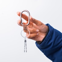 A hand holding a carabiner attached to a keyring with two silver keys. The hand is wearing a blue sleeve, and the background is a plain white surface.