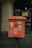A vintage orange mailbox stands on a cobblestone street, adorned with various stickers and weathered paper remnants. The backdrop features the entrance of a shop with a partially visible display and a yellow sign indicating a 30% discount.